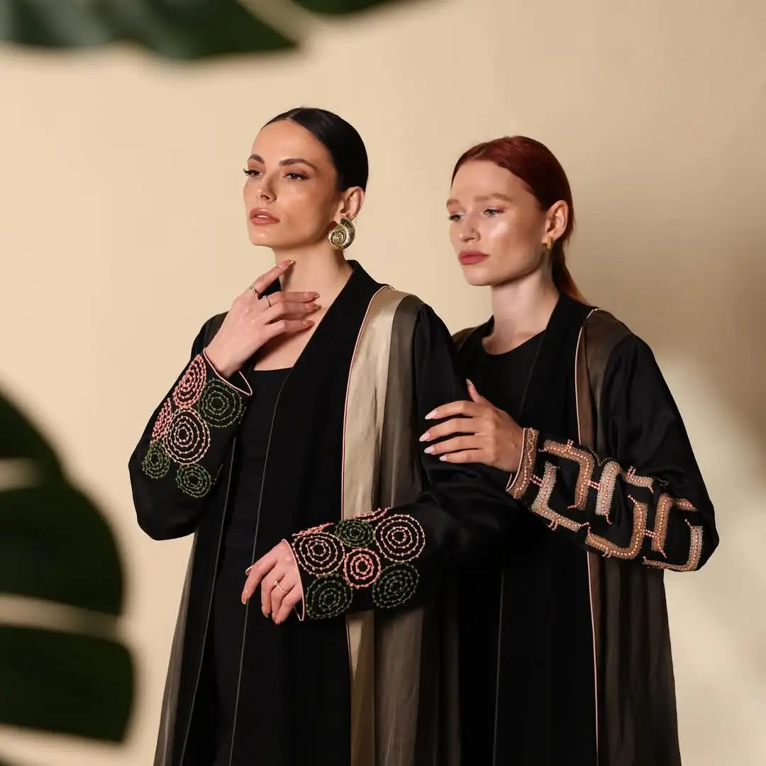 Two women wearing black abayas with intricate designs on a beige background