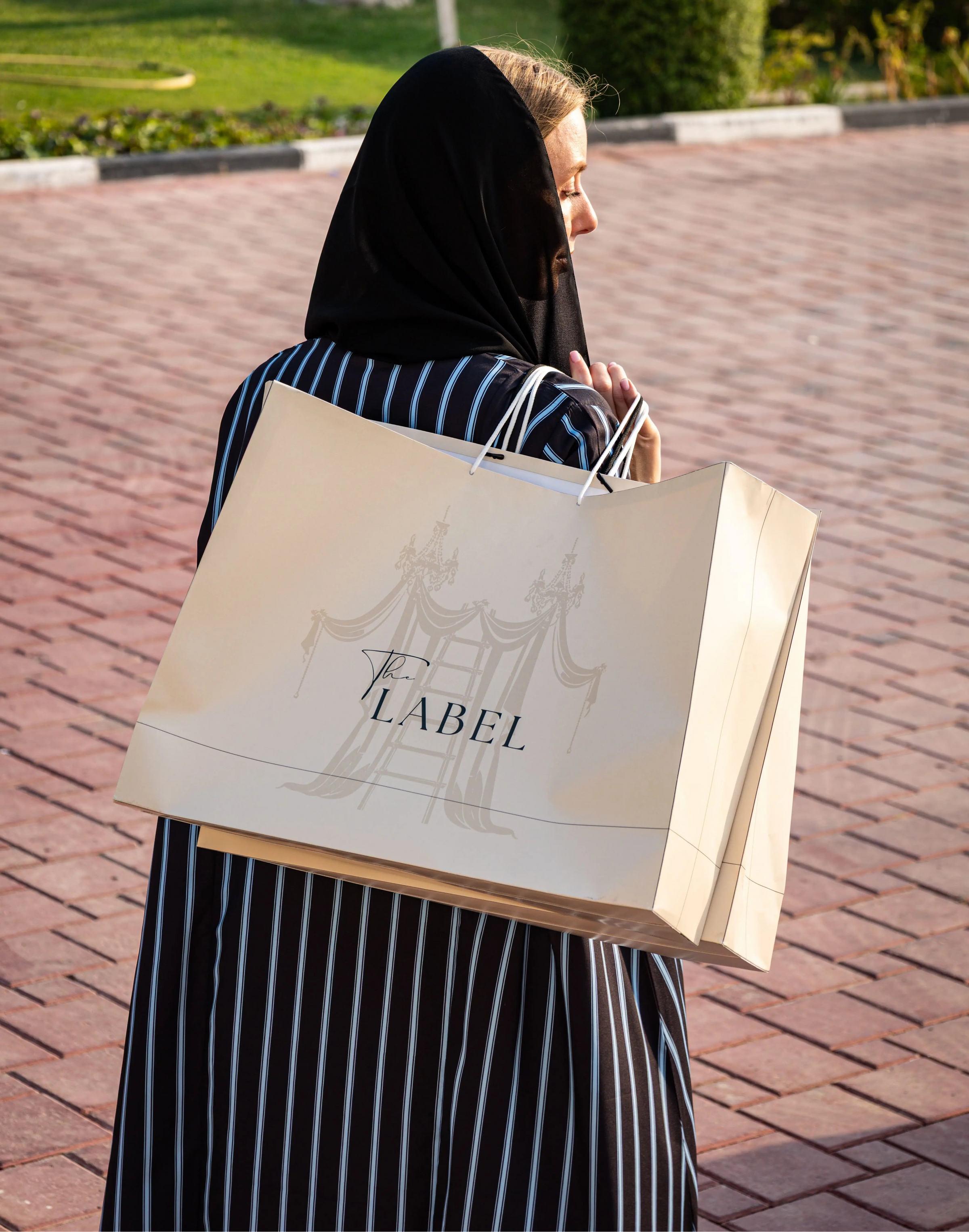 Person holding a shopping bag with 'The Label' on a paved path outdoors